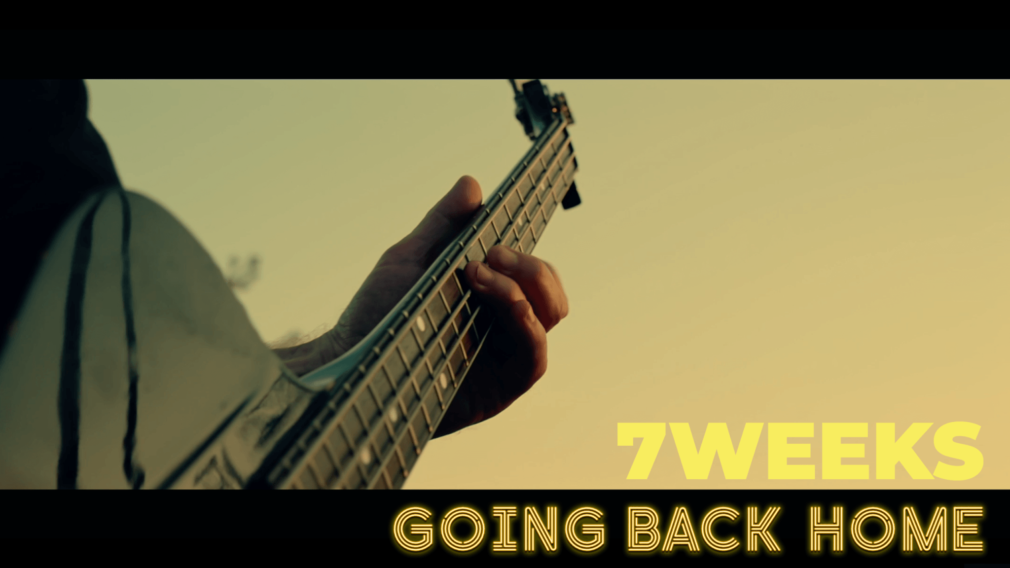 Gros plan sur le manche d'une basse tenue par un musicien en extérieur, avec l'inscription « 7WEEKS - GOING BACK HOME » en jaune